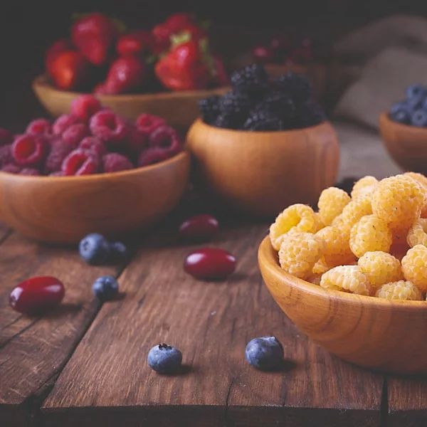 Wooden bowls of fresh berries — raspberries, strawberries, blackberries, blueberries and cloudberries — on a rustic table