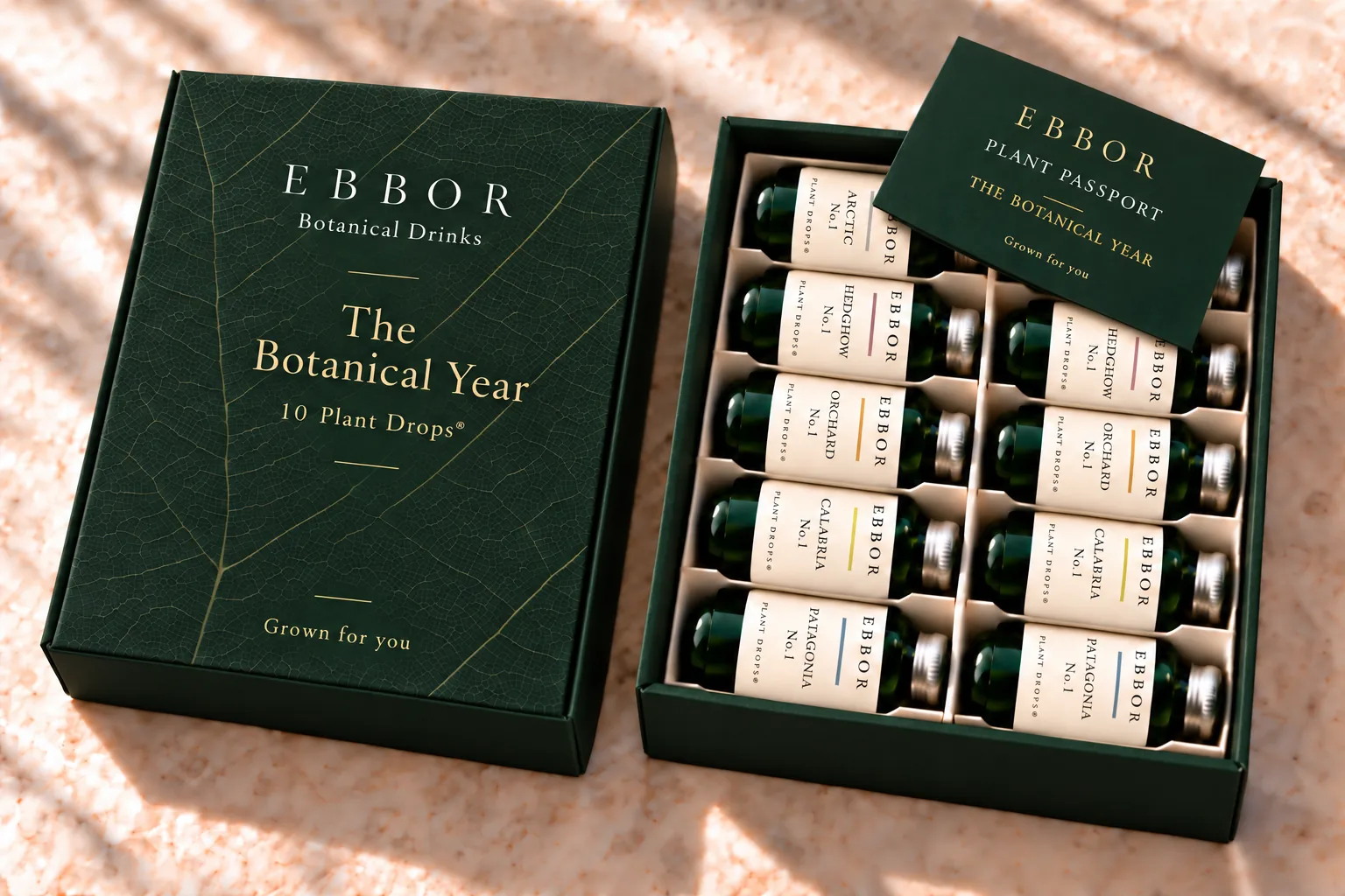 The Botanical Year box: a deep green box printed with a leaf-vein pattern, its lid resting beside an open tray of ten Plant Drop® bottles, with the Plant Passport card lying across the top