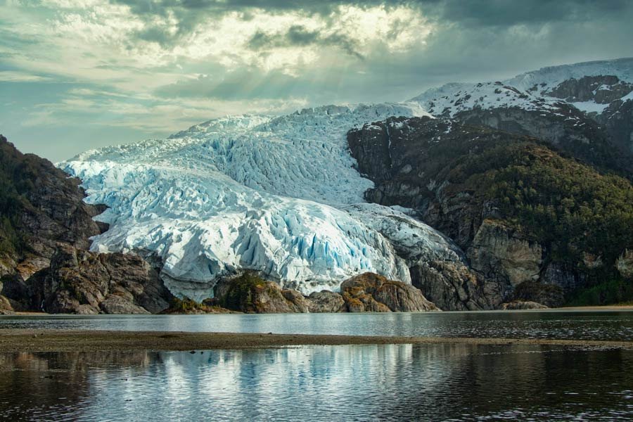 A glacier meeting calm water below steep mountain slopes, cloud breaking above